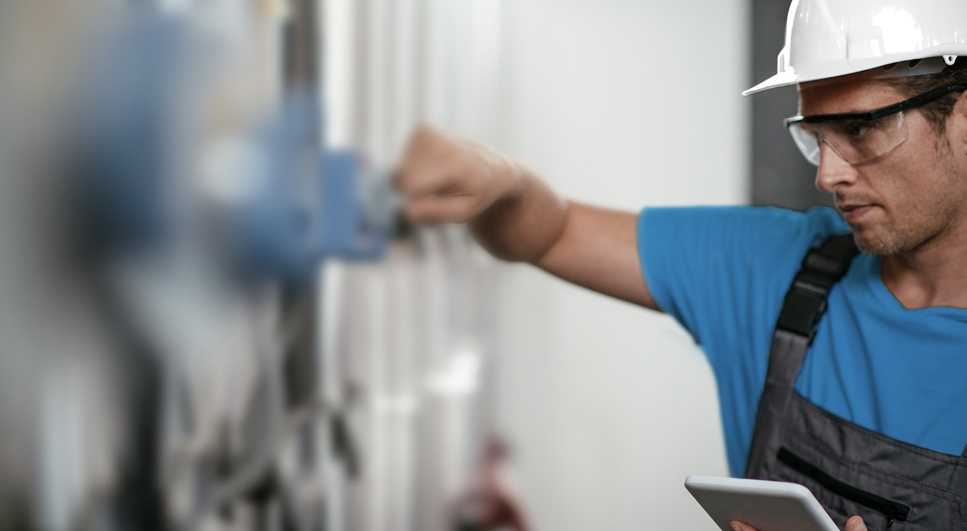A man with a white construction worker's helmet installs a meter. He is holding a tablet in his hand.