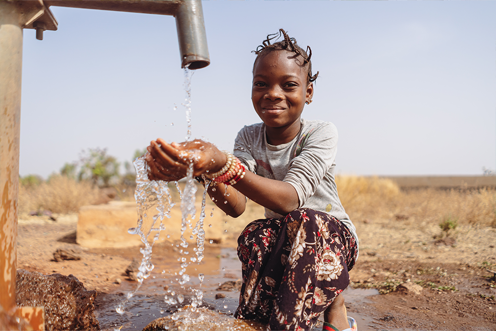 A young girl is crouching in front of a tap from which water is flowing, in a desert-like setting, and is holding her hands under the water.